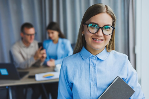 Young business lady Manager woman in classic glasses smiling during working time in office desk with laptop colleagues in background A colleague is in the background selective focus