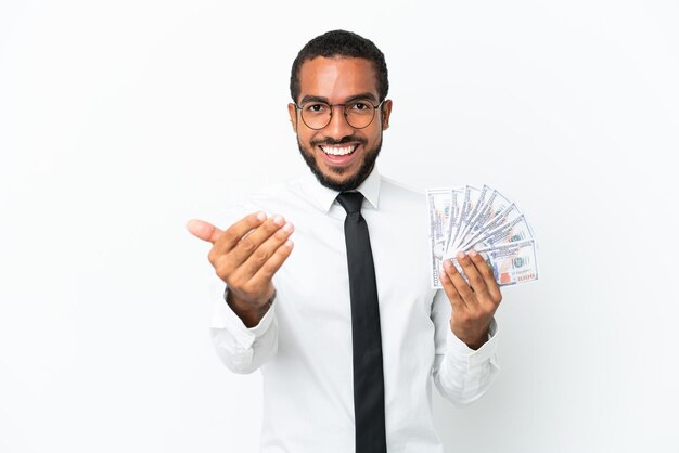 Young business latin man taking a lot of money isolated on white background inviting to come with hand Happy that you came