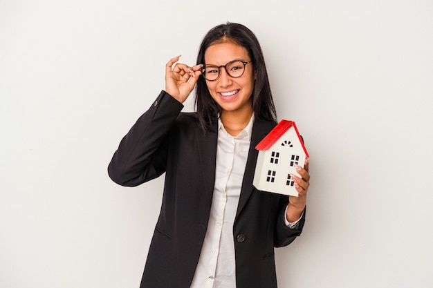 Young business latin woman holding a toy house isolated on white background  excited keeping ok gesture on eye.