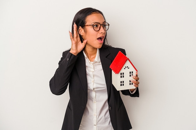 Young business latin woman holding a toy house isolated on white background  trying to listening a gossip.