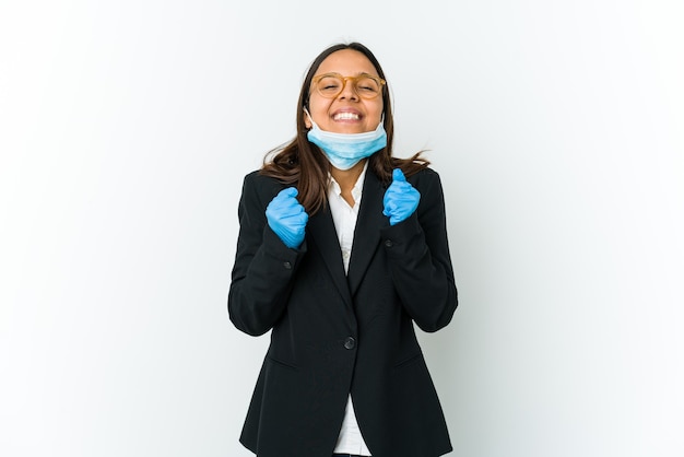 Young business latin woman wearing a mask to protect from covid isolated on white raising fist, feeling happy and successful. Victory concept.