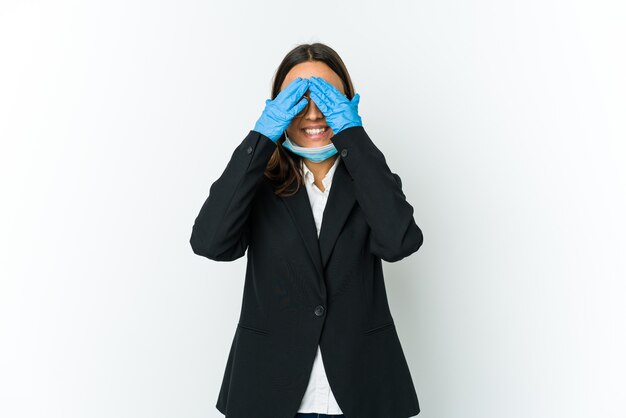 Young business latin woman wearing a mask to protect from covid isolated on white wall covers eyes with hands, smiles broadly waiting for a surprise.