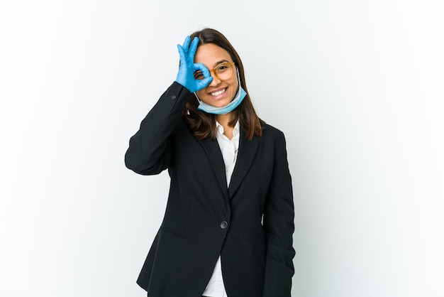 Young business latin woman wearing a mask to protect from covid isolated on white wall excited keeping ok gesture on eye.