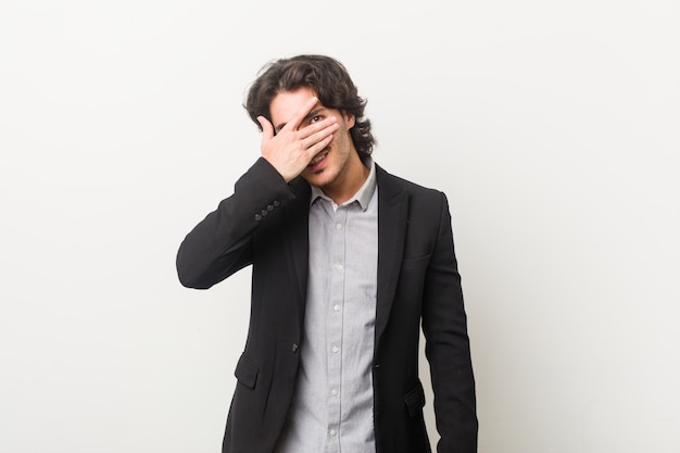 Young business man against a white wall blink at the camera through fingers, embarrassed covering face.