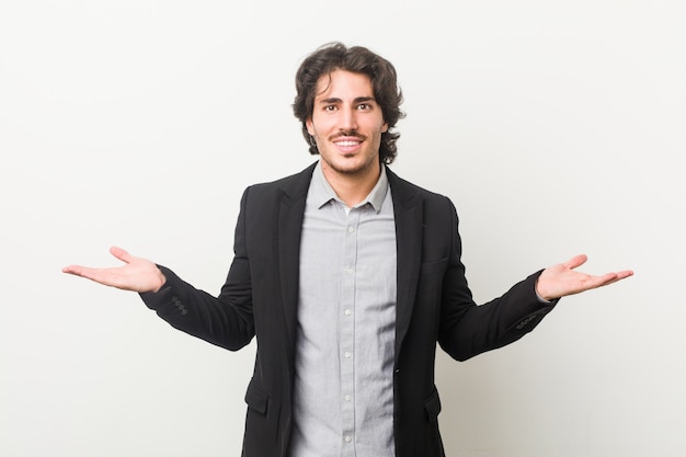 Young business man against a white wall makes scale with arms, feels happy and confident.