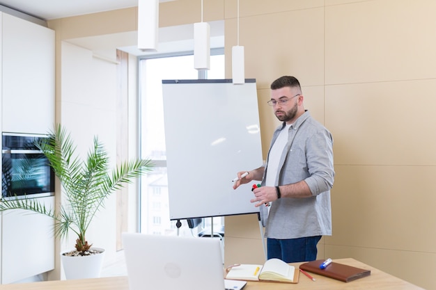 Giovane uomo d'affari in abiti casual riunione online presentazione o formazione utilizzando una webcam portatile e una lavagna a fogli mobili con pennarelli