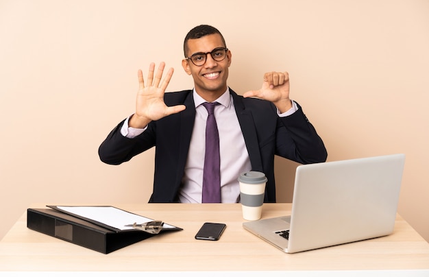 Молодой деловой человек в своем офисе с ноутбуком и другими документами, считая шесть с пальцами
