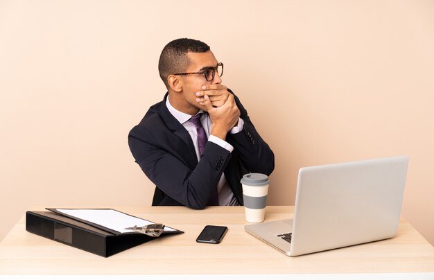 Молодой деловой человек в своем офисе с ноутбуком и другими документами, охватывающими рот и глядя в сторону