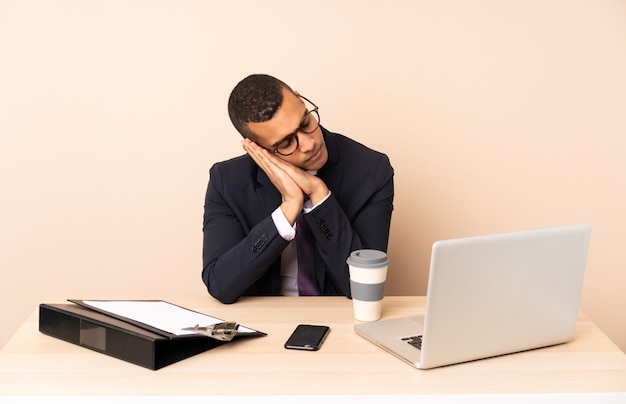 Il giovane uomo di affari nel suo ufficio con un computer portatile e altri documenti che fanno il sonno gesturing nell'espressione dorable