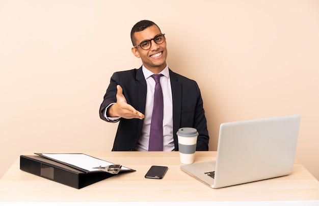 Young business man in his office with a laptop and other documents shaking hands for closing a good deal