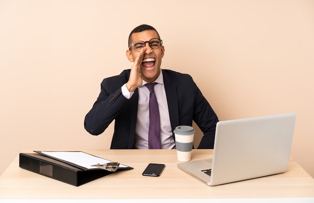 Young business man in his office with a laptop and other documents shouting with mouth wide open