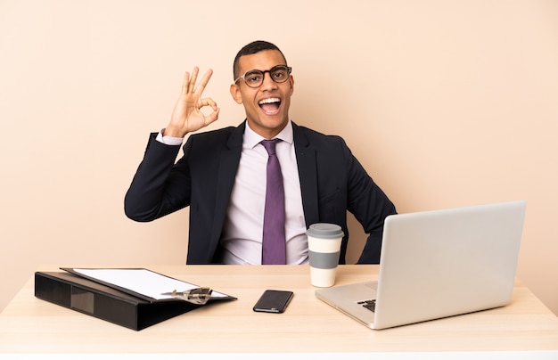 Young business man in his office with a laptop and other documents surprised and showing ok sign