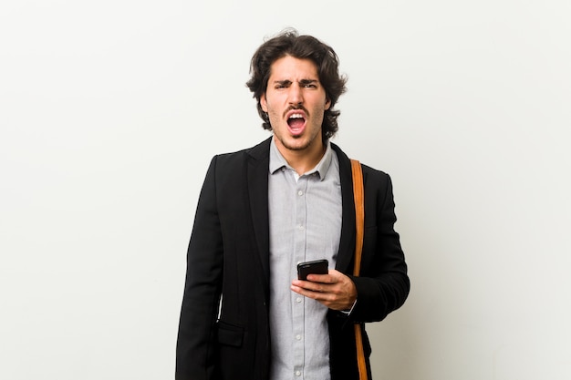 Young business man holding a phone screaming very angry and aggressive.