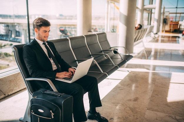 Young business man sitting on the computer with the suitcase at the airport waiting for the flight xAxA