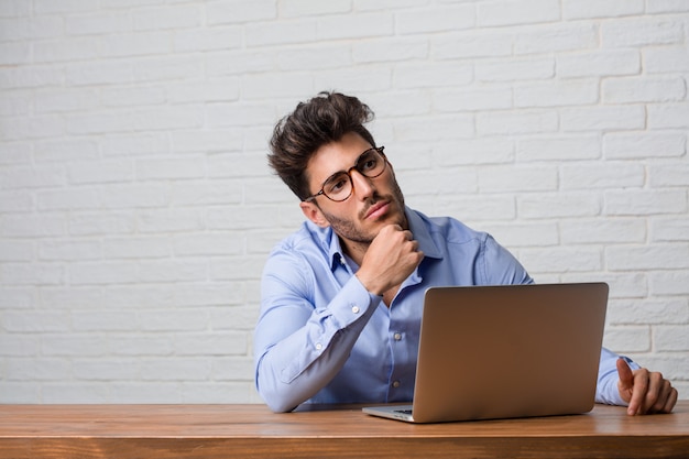 Giovane uomo di affari che si siede e che lavora ad un computer portatile che pensa e che osserva in su