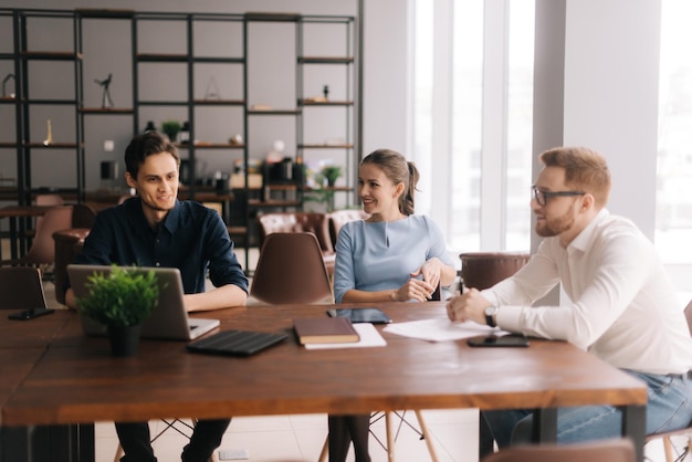 Young business man tells his colleagues about the project in modern office meeting against large panoramic window Young man and woman listen to the speaker Concept of office life