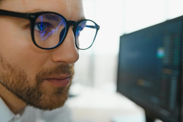 Young business man trader wearing glasses looking at computer screen with trading charts reflecting in eyeglasses watching stock trading market financial data growth concept close up