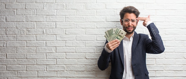 Young business man wearing a suit against a white bricks wall making a suicide gesture