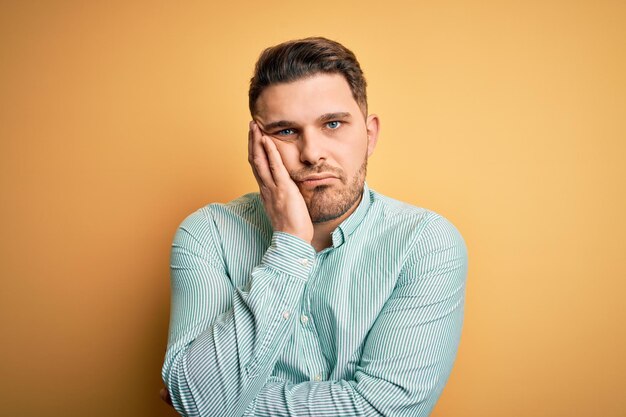 Young business man with blue eyes wearing elegant green shirt over yellow background thinking looking tired and bored with depression problems with crossed arms