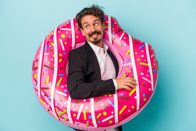 Young business man with inflatable donut isolated on blue background looks aside smiling, cheerful and pleasant.