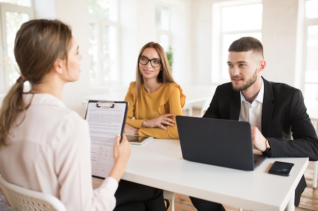 Young business man with laptop and business woman in eyeglasses talking with applicant about work Young smiling employers spending job interview in modern office