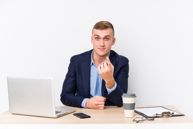 Young business man with a laptop pointing with finger at you as if inviting come closer.