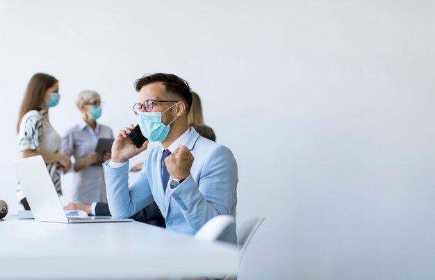 Il giovane uomo di affari con la mascherina protettiva medica lavora ad un computer portatile e usando il telefono cellulare nell'ufficio