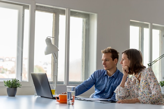 Young business man and woman sitting at a white office desk working together using laptop computer.