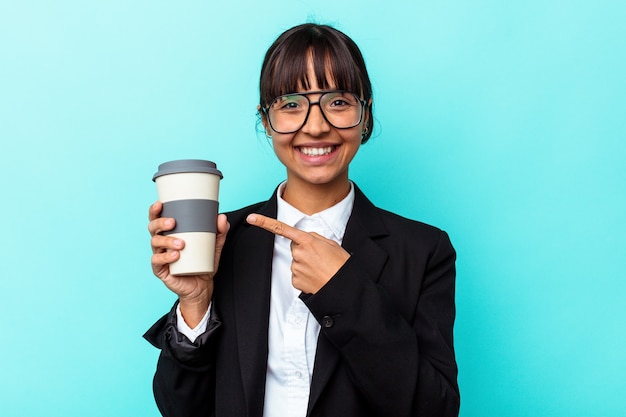 青い背景に分離されたコーヒーを持って笑顔で脇を指して、空白のスペースで何かを示す若いビジネス混血の女性。