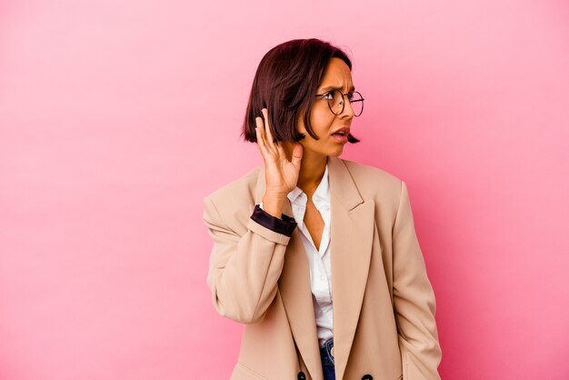 Young business mixed race woman isolated on pink wall trying to listening a gossip