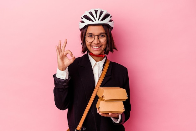 Young business mixed race woman wearing bike helmet holding burger isolated on pink background cheerful and confident showing ok gesture.