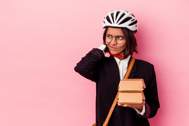 Giovane donna di razza mista d'affari che indossa un casco da bici che tiene hamburger isolato su sfondo rosa toccando la parte posteriore della testa, pensando e facendo una scelta.