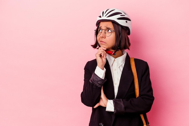 분홍색 배경에 격리된 자전거 헬멧을 쓴 젊은 비즈니스 혼혈 여성이 의심스럽고 회의적인 표정으로 옆을 바라보고 있습니다.
