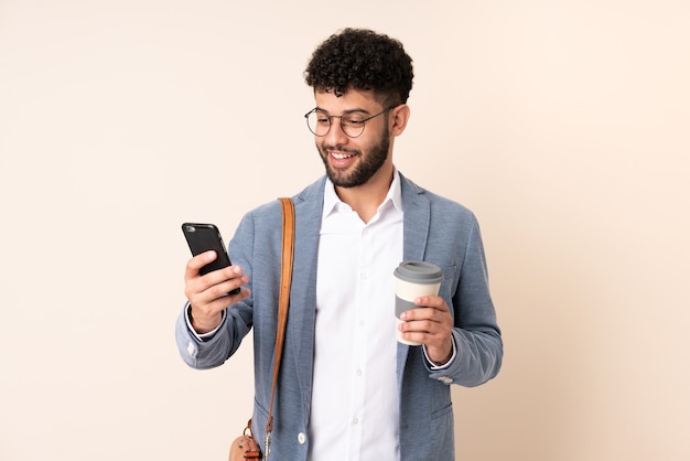 Young business Moroccan man isolated on beige background holding coffee to take away and a mobile