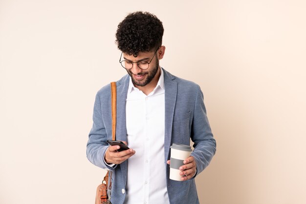 Young business Moroccan man isolated on beige wall holding coffee to take away and a mobile