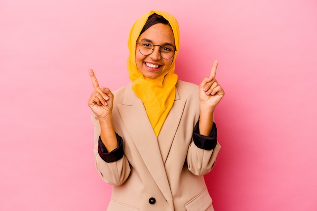 Young business muslim woman on pink indicates with both fore fingers up showing a blank space.