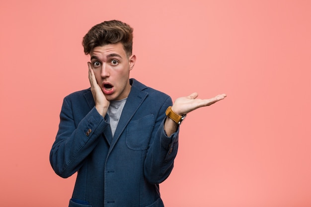 Young business natural man holds copy  on a palm, keep hand over cheek. Amazed and delighted.