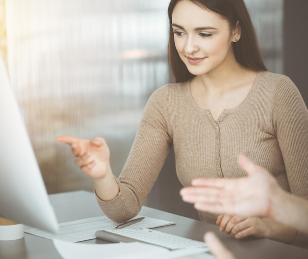 Young business people are talking to each other, while sitting at the desk in sunny office. Businesswoman is explaining something to her colleague.