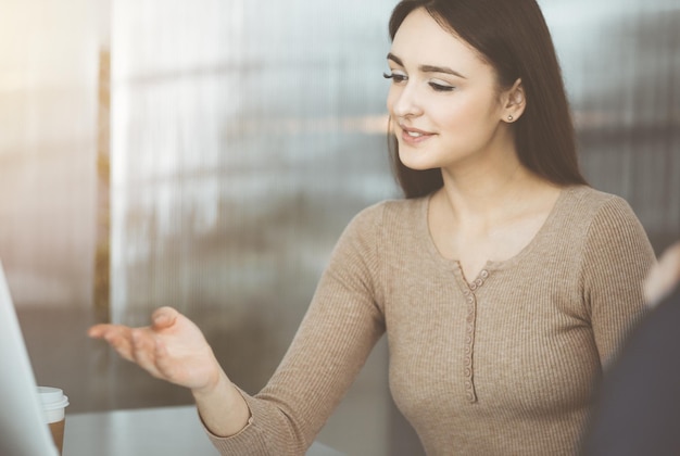 若いビジネスマンは、日当たりの良いオフィスの机に座りながら、お互いに話し合っています。女性実業家が同僚に何かを説明している