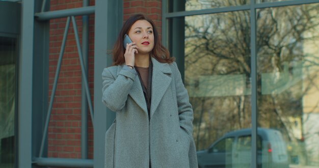 Young business woman in a coat is talking on phone outside near modern office building