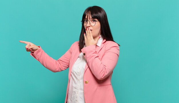 Young business woman feeling happy, shocked and surprised, covering mouth with hand and pointing to lateral copy space