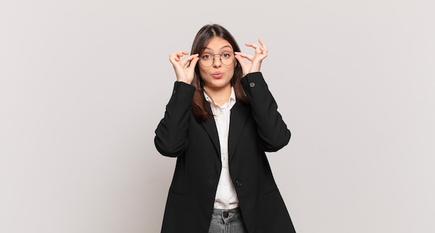 Young business woman feeling shocked, amazed and surprised, holding glasses with astonished, disbelieving look
