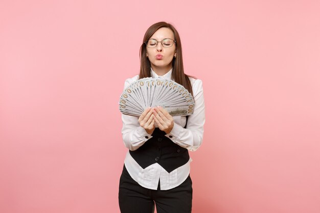 Young business woman in glasses holding bundle lots of dollars, cash money blowing lips kissing isolated on pink background. Lady boss. Achievement career wealth concept. Copy space for advertisement.