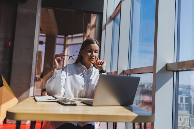 Young business woman in glasses speaks on the phone in the office Business woman texting on the phone and working on a laptop A young business woman is sitting at a table in a cafe Smiling woman