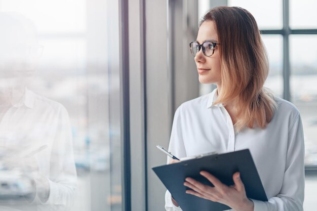 Young business woman holding folder of documents and standing at the panoramic office window