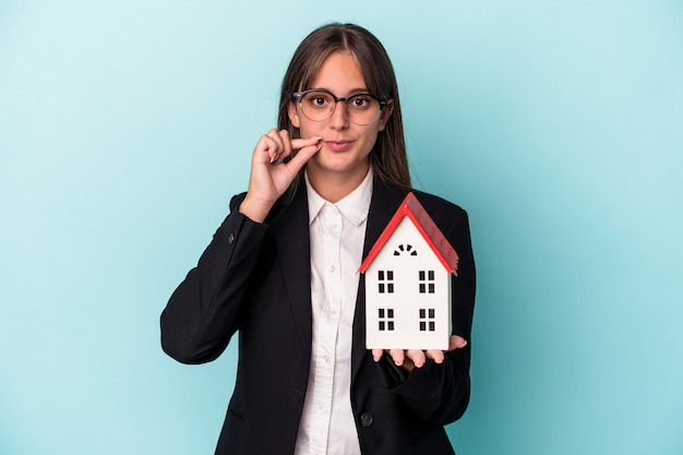 Young business woman holding a toy home isolated on blue background with fingers on lips keeping a secret.