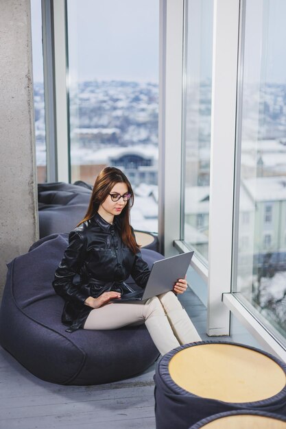 젊은 비즈니스 여성이 노트북 작업을 하는 동안 앉아 있습니다. 현대적인 성공적인 여성의 개념 현대적인 작업 열린 공간에서 의자에 앉아 안경을 쓴 젊은 심각한 매력적인 소녀