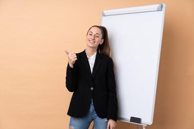 Young business woman over isolated wall giving a presentation on white board and with thumbs up