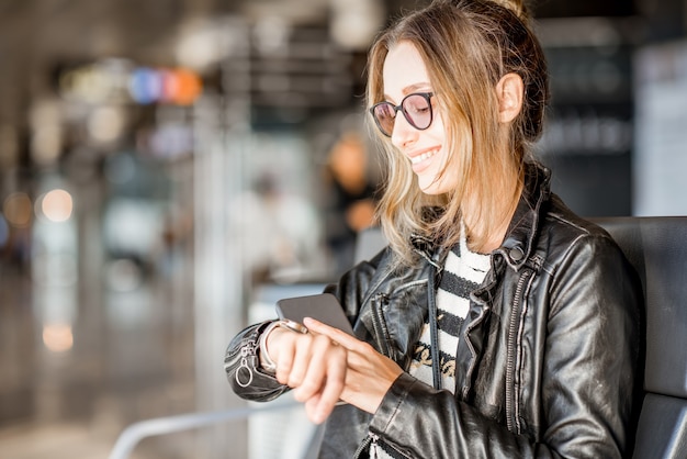 Young business woman in leather jacket and pants sitting with phone at the departure hall of the airport waiting for the flight