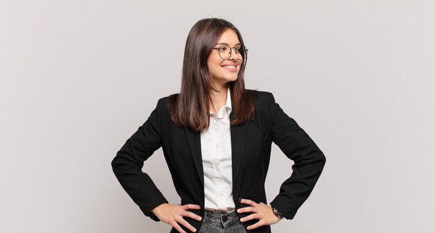 Young business woman looking happy, cheerful and confident, smiling proudly and looking to side with both hands on hips